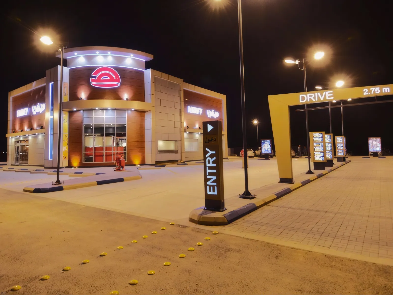A Herfy drive-thru at night, with lit digital menu pylons along the drive lane
