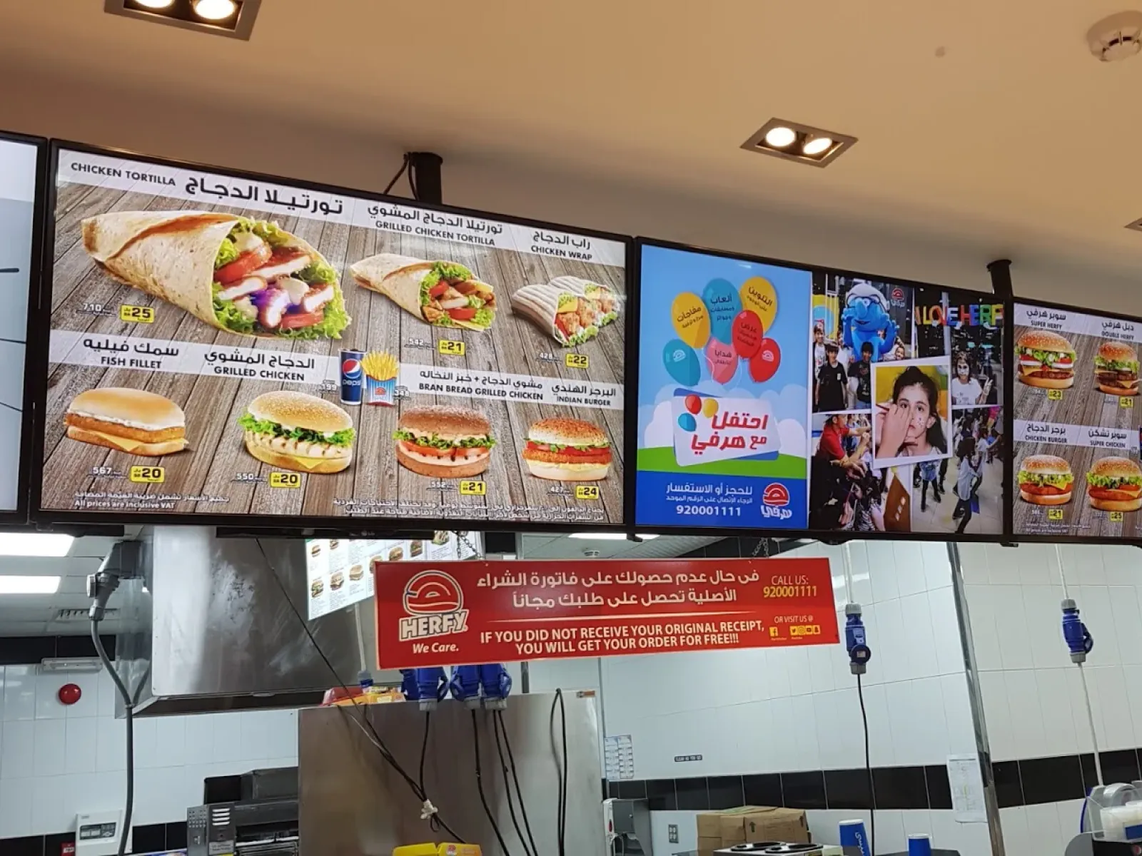 Bilingual Arabic and English menuboards above a Herfy counter, next to a screen advertising kids' parties