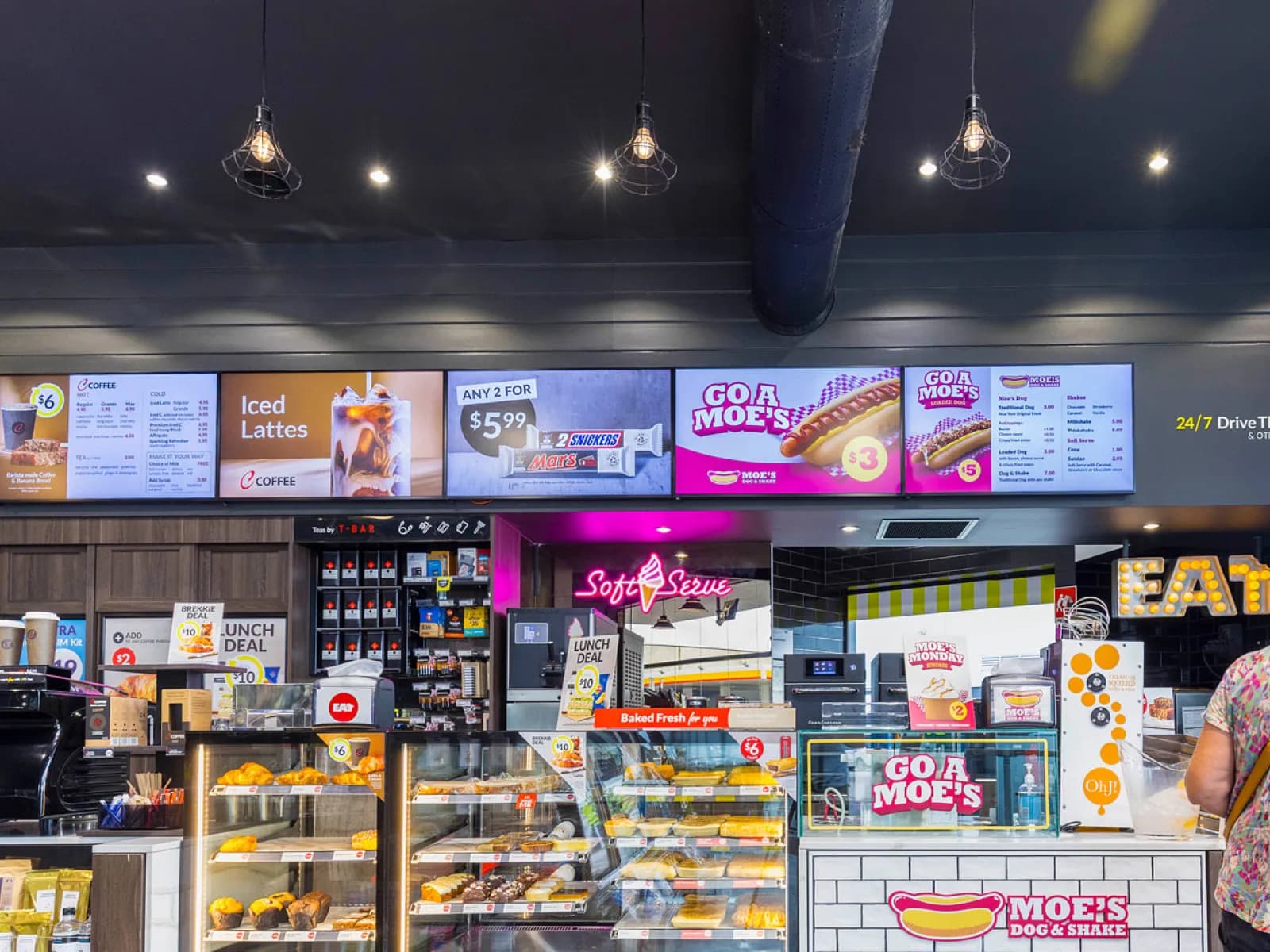 A row of menu screens above the counter inside an OTR store, for coffee, snacks and Moe's Dog & Shake