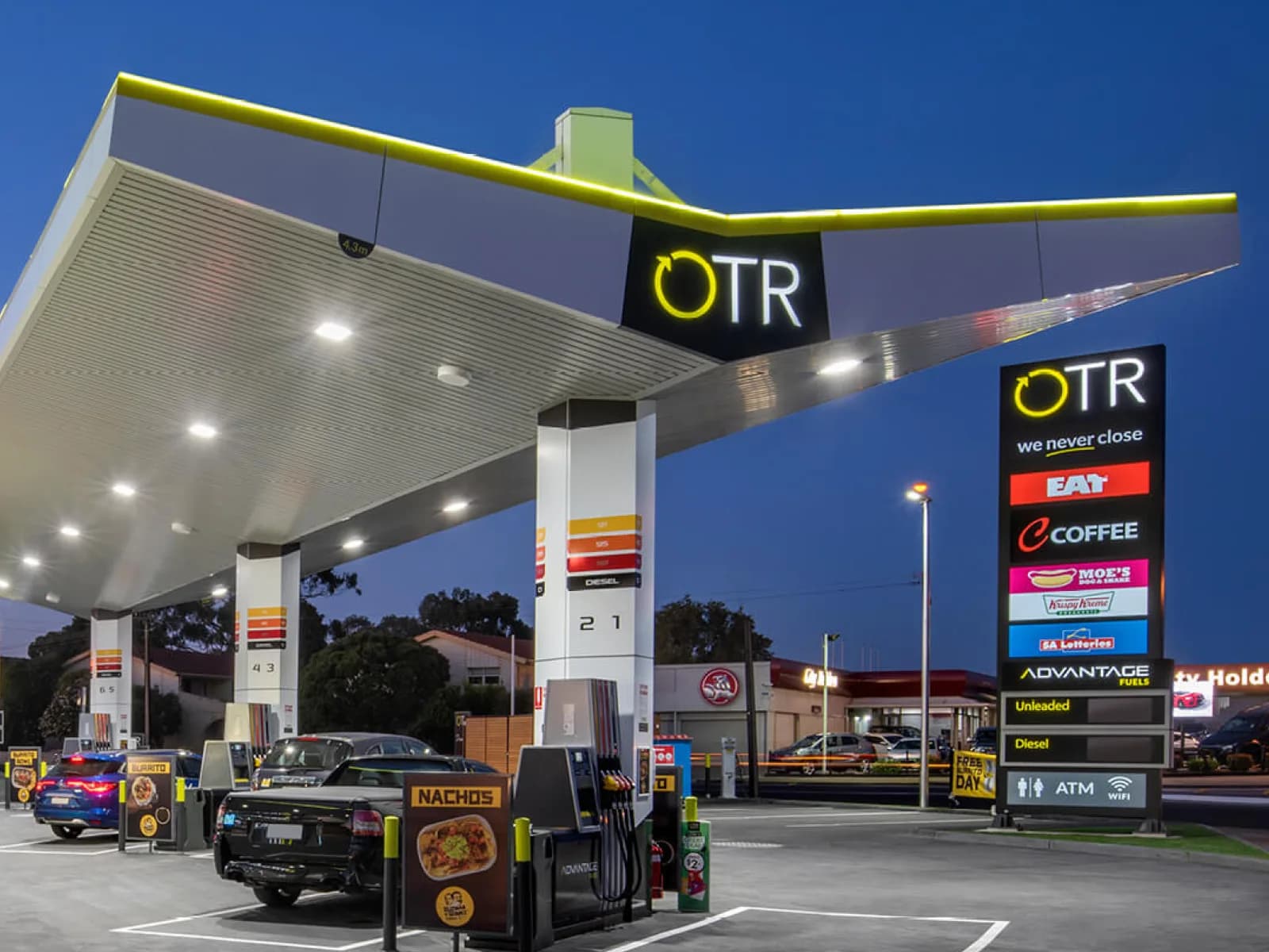An OTR forecourt at dusk, with the canopy lit and the fuel price pylon beside the road