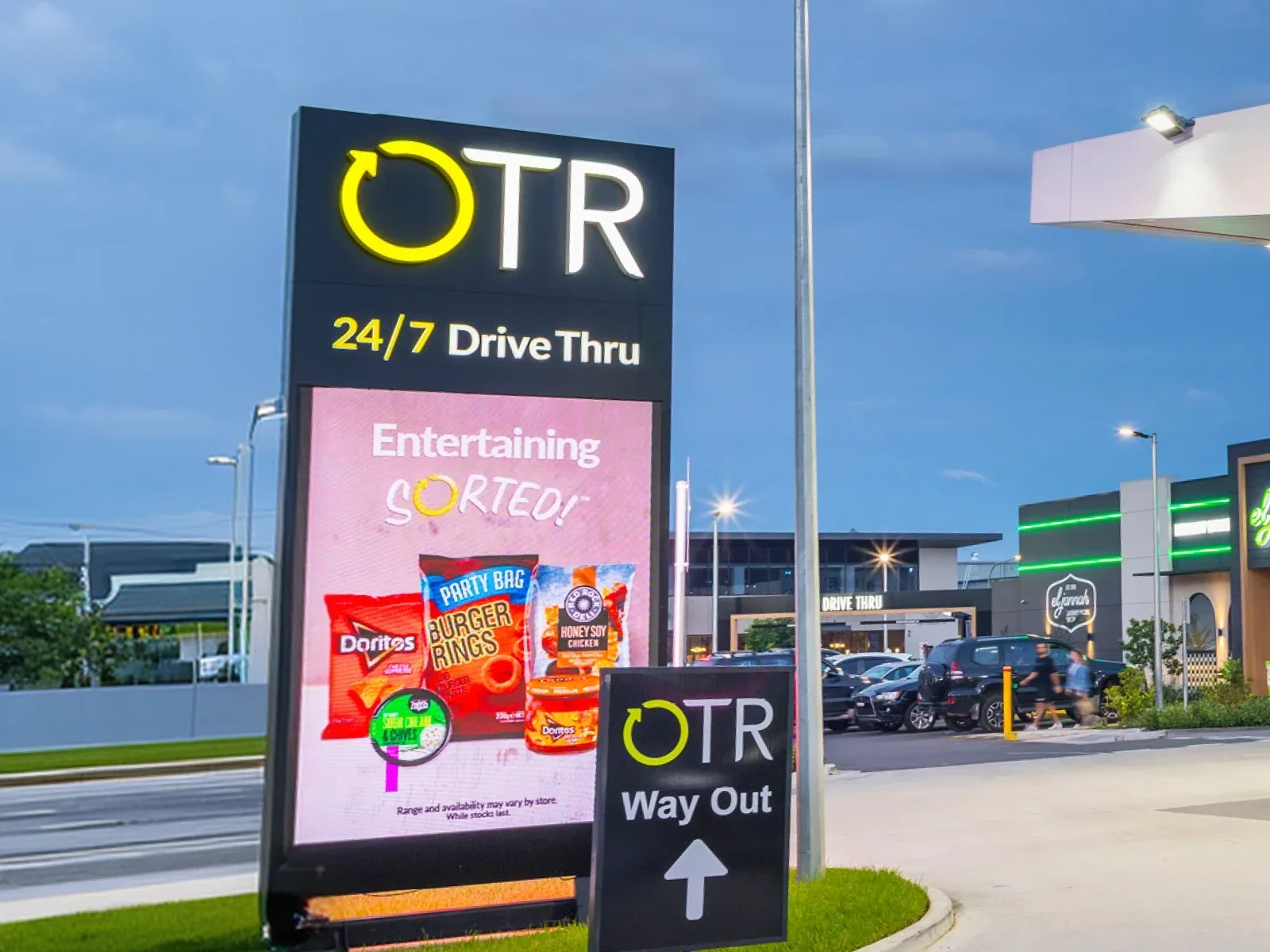 An OTR 24/7 Drive Thru pylon by the road, with a promotion playing on its digital screen