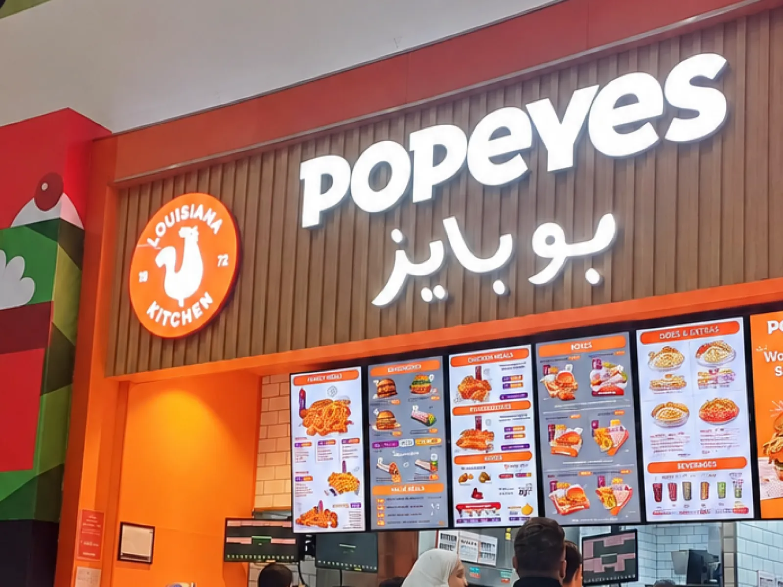 A Popeyes counter in a mall, with a row of portrait menuboards under the lit Popeyes sign in English and Arabic