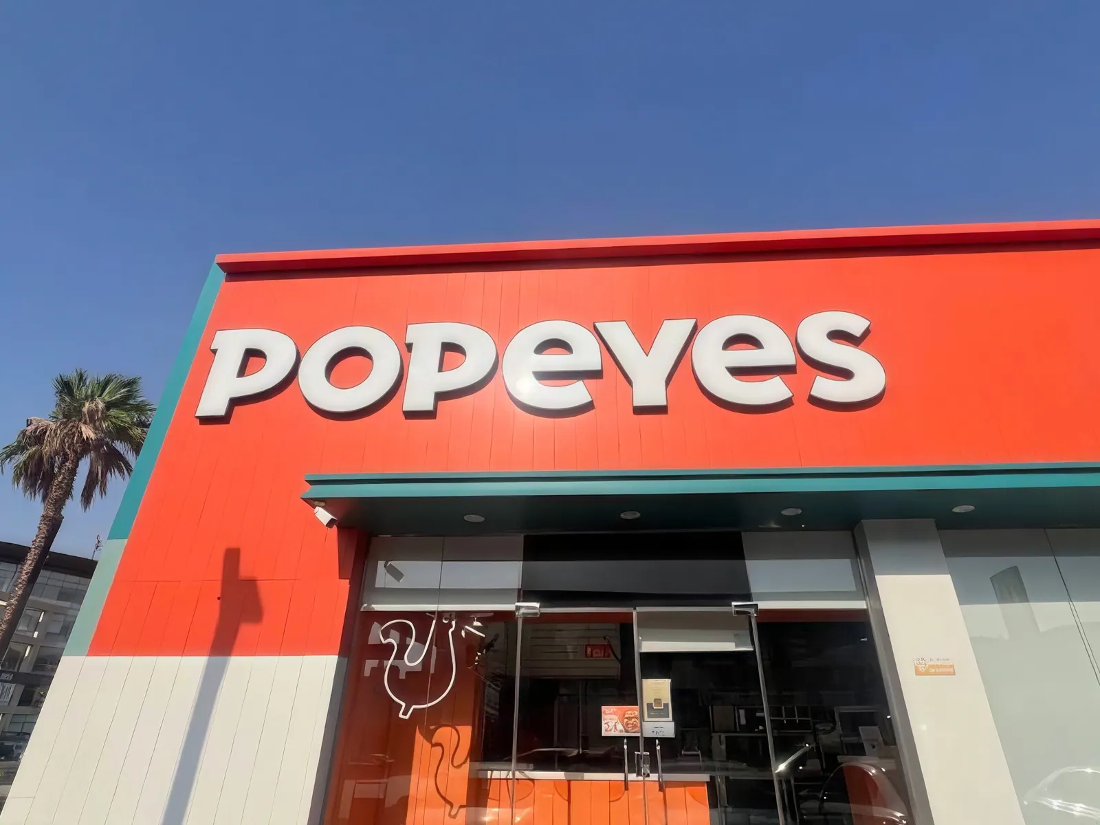 The front of a Popeyes store, with the white Popeyes lettering on an orange facade under a clear sky