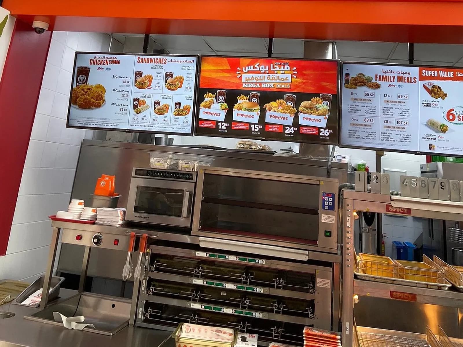 Three landscape menuboards above a Popeyes kitchen, showing chicken combos, Mega Box deals and family meals priced in Saudi riyals