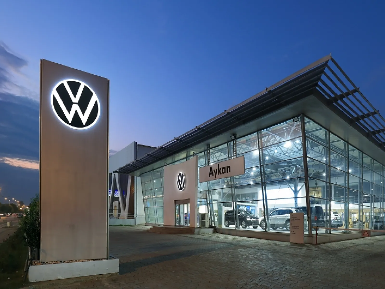 A Volkswagen dealership at dusk, with a lit VW pylon outside the glass showroom
