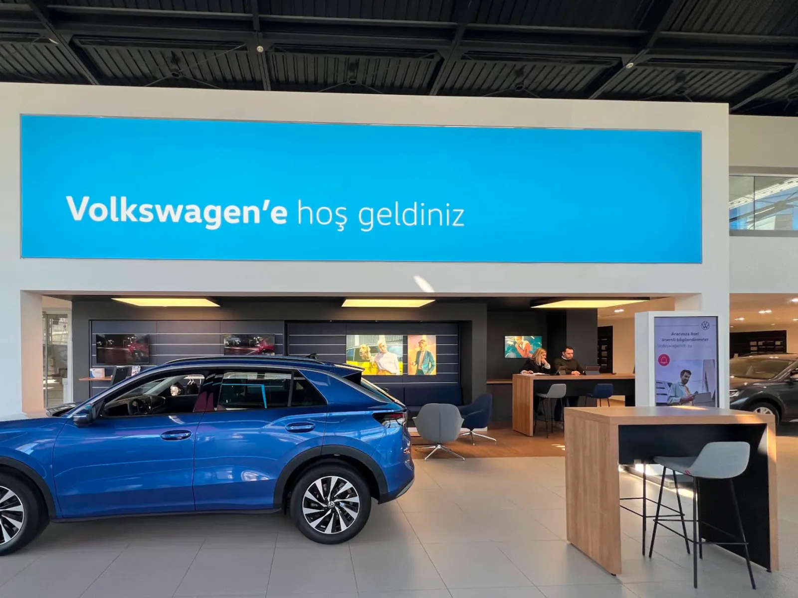 A Volkswagen showroom with a wide welcome screen above the customer lounge, a blue SUV in front and a portrait information kiosk to the right