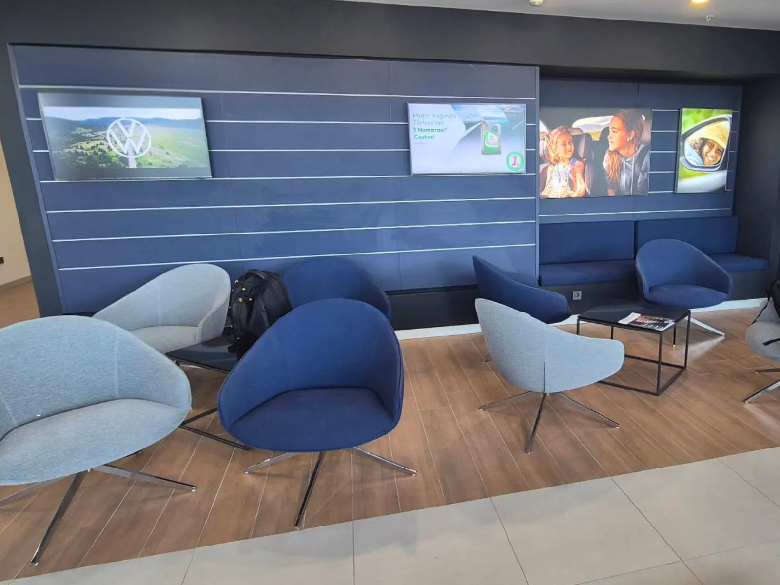 A Volkswagen service lounge with blue armchairs and wall screens showing Volkswagen brand content and a Castrol motor oil ad
